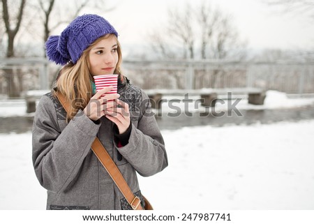 Woman in grey coat holding a cup of  hot coffee with milk. Lonely girl stands in winter snowy city park
