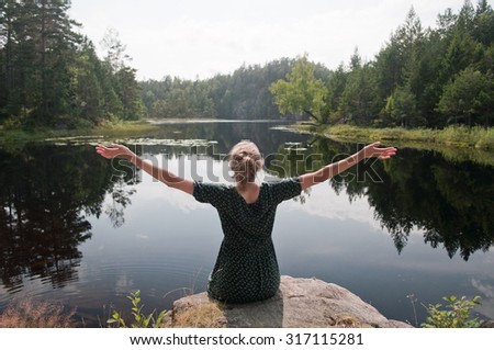 Traveler woman raising hands looks at the landscape of summer Norway. Forest, lake and sky