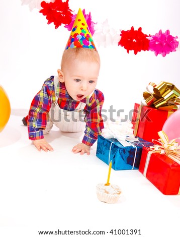 Baby in a party hat looks at the birthday cake with surprise