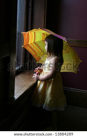 Little girl with yellow umbrella, looking out a large window.