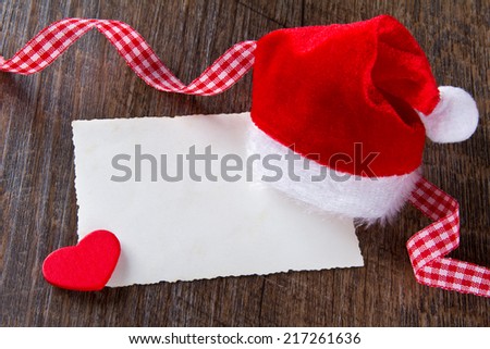 Father Christmas cap with yellowed card, red heart and checkered ribbon on dark wood, copy space