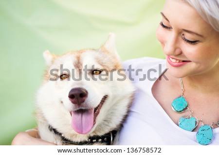 A youthful woman is smiling as she holds a young adult dog. The dog is a Siberian Husky/Alaskan Malamute of red and white coloring. They lovingly and comfortably sit by a green backdrop.