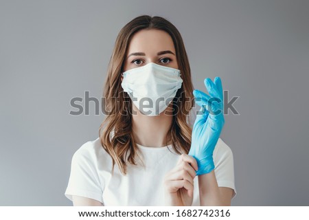 Young woman patient in a medical mask puts on protective surgical sterile gloves on her arm, isolated on gray background, protection against coronovirus ストックフォト © 