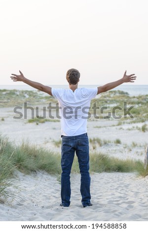 Young man standing on dune front of sea, back to the camera with hands up.