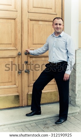 A well dressed man stands in front of a large wooden door. Keep the door handle. Ready to go in it.