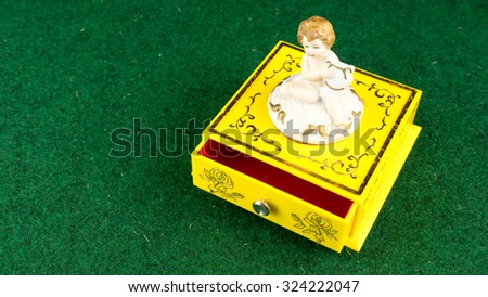 Yellow colour winding music or jewellery box case with white cute angelic cupid statue playing musical item on top surrounded by green grass background. Slightly defocused and close up. Copy space.