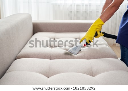 Close-up of housekeeper holding modern washing vacuum cleaner and cleaning dirty sofa with professionally detergent. Professional springclean at home concept