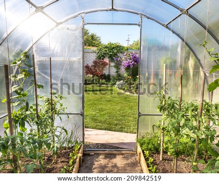 Plants growing in a translucent domed greenhouse or tunnel with a view from inside through the open door onto a pretty formal garden