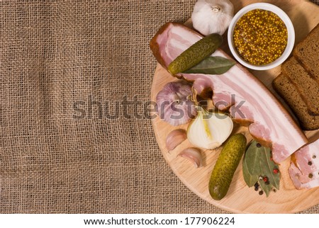 Still Life: Bacon, Dijon Mustard, Bread, Garlic, Onion, Pickles on Round Wooden Board and Sackcloth Background