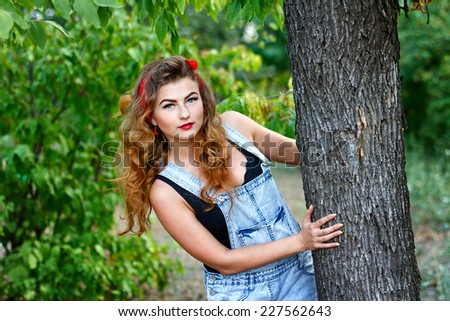 Beautiful pin-up girl in denim overalls and a a red kerchief hiding behind a tree