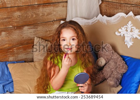 Little girl with long curly hair holds in hands mirror and lip gloss and makeup