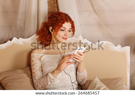 Young attractive redhead curly girl listening to music on headphones, holding a smart-phone. Girl sitting on the bed in bedroom. The concept of youth and technology.