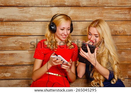 Two sisters, blonde, listening to music on headphones. Girls having fun and looking at the screen cell, holding a tablet. Sisters sitting in the bedroom.