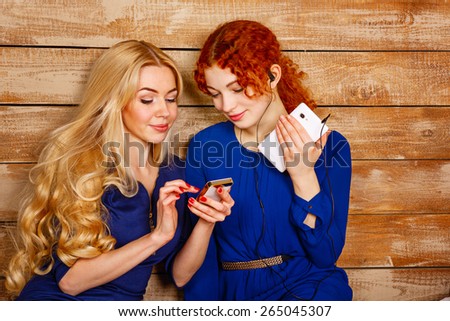 Two sisters, a blonde and a redhead, listening to music on headphones. Girl shows the latest news on the screen of your smartphone. Sisters sitting in the bedroom.