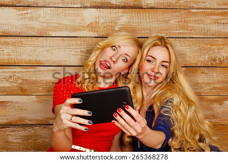 Young blonde sisters make fun group selfie, holding a tablet computer. A girl shows tongue. The concept of youth and technology.