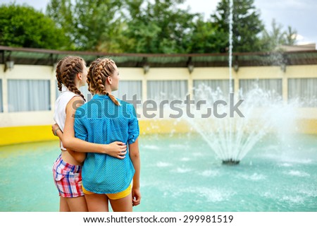 Friendly hug at the fountain in summer park. Girls dressed in shorts and a shirt. On summer vacation. The concept of true friendship.