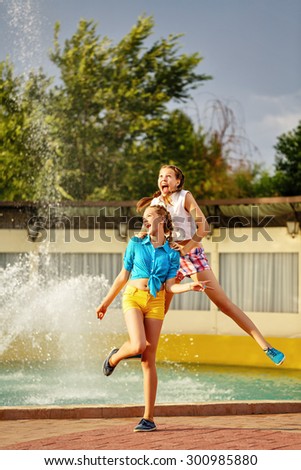 Girl jumping, holding the shoulders of her best friend, near the fountain in the summer park. Girls dressed in shorts and a shirt. On summer vacation. The concept of true friendship.