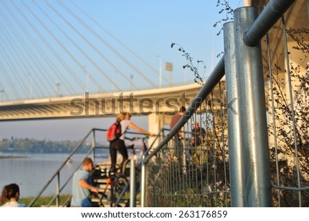 Defocused cyclists on their bicycles shot from behind, passing under a bridge, on a sunny afternoon. Focus on the iron fence.