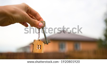 Hand with a key and a wooden key ring-house. Background of fence and cottage. Building, project, moving to a new home, mortgage, rent and purchase real estate. To open the door. Copy space