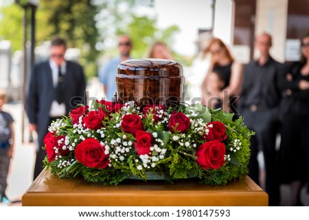 Funerary urn with ashes of dead and flowers at funeral. Burial urn decorated with flowers and people mourning in background at memorial service, sad and grieving last farewell to deceased person.