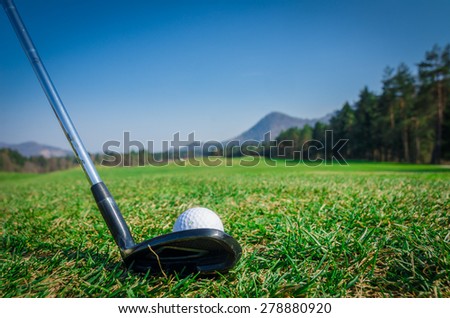 Chipping a golf ball onto the green with driver golf club. Green grass with forrest and mountains in the background. Soft focus or shallow depth of field. Back view