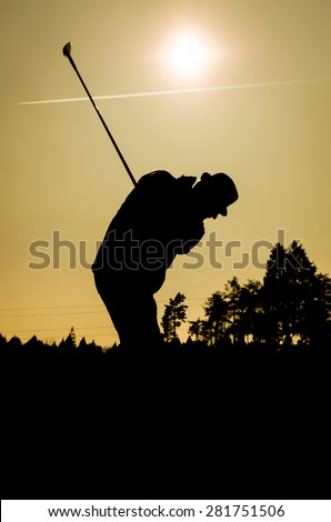 Silhouette of old man playing golf. Senior citizen is wearing a hat and swinging the club with sun and airplane contrails in the background. Also forest and trees in evening or morning dusk or dawn.