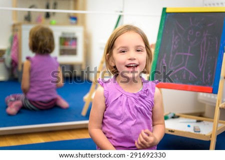 Little child is drawing with pieces of color chalk on the chalk board. Girl is expressing creativity and looking at the camera, smiling in a nursery, classroom or playroom. Concept of learning
