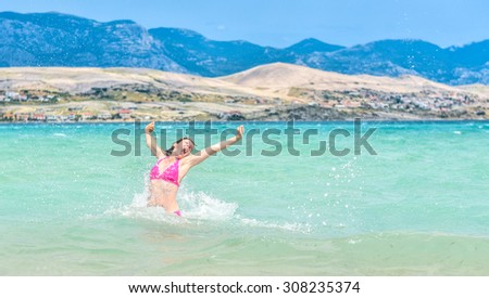 Woman is jumping out of water smiling, celebrating healthy lifestyle. Arms stretched in blue sea with island in the background.