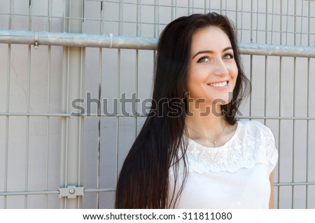 Half-length portrait of beautiful smiling young brunette woman with long hair wearing in a white shirt and denim shorts on metal mesh background