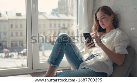 Close up pretty young woman sitting on a windowsill at home and texting on her phone communication female looking message cellphone cheerful smile use internet modern smartphone slow motion Foto d'archivio © 