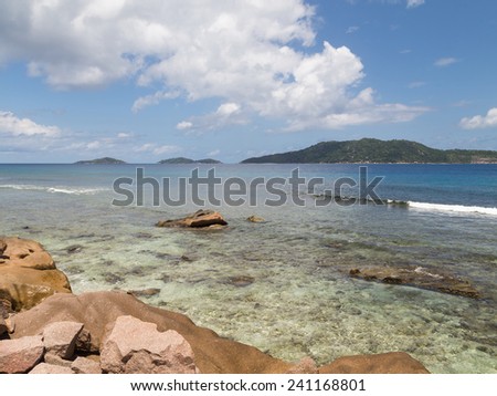 tropical landscape with large granite stones, clean ocean with waves and green mountains on the island of La Digue, Seychelles