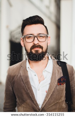 young guy with a beard and mustache with glasses in a fashionable jacket posing on the street
