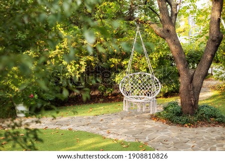 Comfortable hanging wicker white chair in summer garden. Cozy hygge place for weekend relax in garden. Hammock chair in boho style hanging on tree. Cozy exterior backyard. Concept of rest outdoor. 
