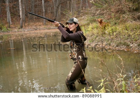 Woman Duck Hunter wearing Camo Waders with Rifle in Pond and Dog in Background