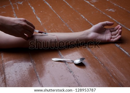 The man's hand lies on a floor, the syringe, a spoon nearby lie