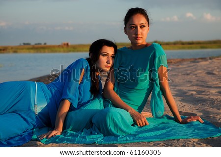 Two beautiful girls lie on a beach and look a sunset, one in a green dress, another in dark blue