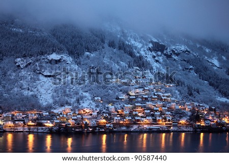 Small town with cottages, on a mountain side with pines and rock, snow covered, over water. Winter, evening time. With cloud in upper part, and reflections in the water.
