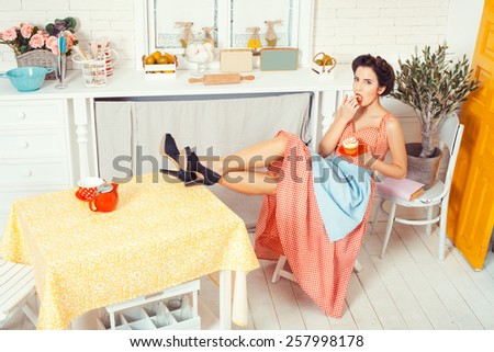 Pin-up girl style. The view from the top. The girl in the kitchen sitting on a chair with his feet on the table. She licks cream muffin.