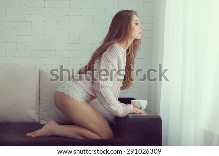 Woman kneels on the couch and looks out the window. Photo toned in vanilla color.
