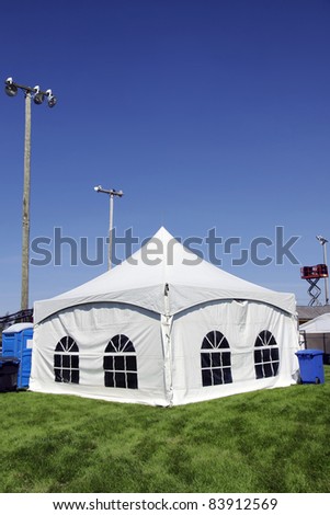 Celebration or event: White tent on soccer field with lighting ready for guests in case of rain with sound system on lift in the background, portable toilet and bins for disposal.