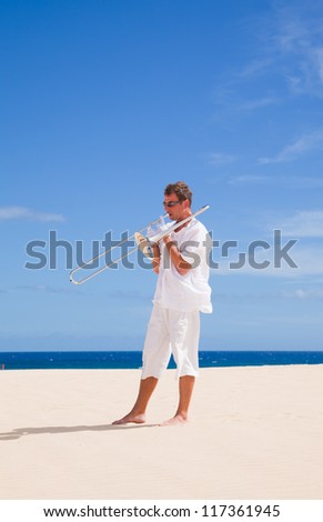 tanned man in white playing silver trombone in the dunes by the ocean