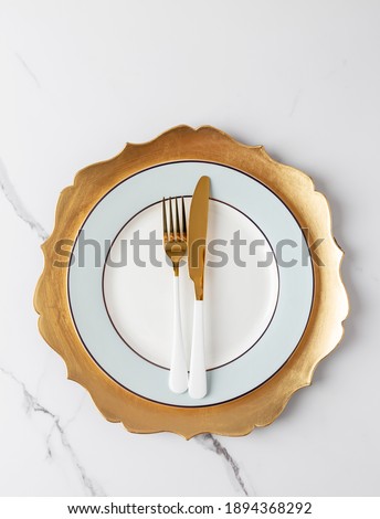 Simple table set with golden and white tableware on white marble. Gold fork and knife at white blue plate with gold dish