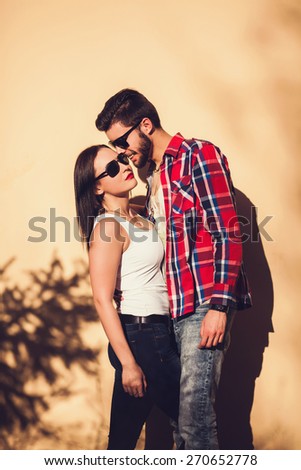 Young fashion elegant stylish couple posing on streets of european city. Sensual brunette vogue girl with handsome hipster man posing near the wall.