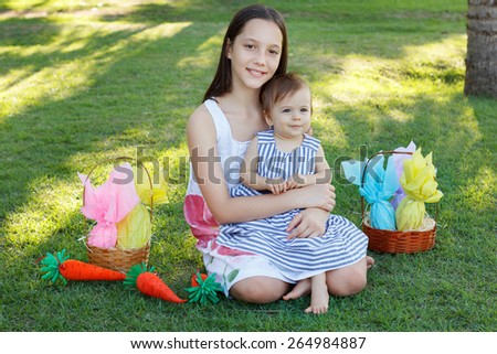 Smiling beautiful teen girl holding sister -  little baby girl on green grass in park with wicker basket with chocolate eggs for Easter. Selective focus
