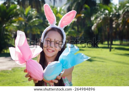 Smiling cute teen girl with rabbit ears holding Easter chocolate eggs in colorful paper in park. Selective focus on face