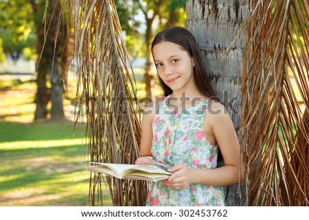 Cute smiling teen girl reading book and looking to camera near palm tree, green grass in park. Selective focus
