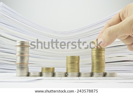 Man putting gold coins on bar chart from stack gold and silver coins with pile of paperwork as background.
