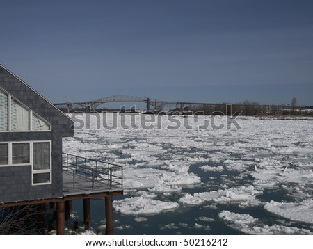 Spring ice flow down the Saint Clair river. There is a house on the rivers edge and the Blue Water Bridge in the background.