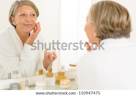 Senior woman looking in bathroom mirror applying anti-wrinkles face cream