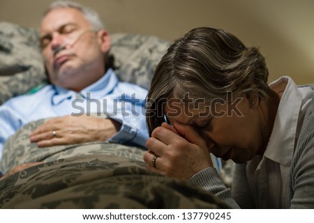Senior woman praying for sick man sleeping in hospital bed